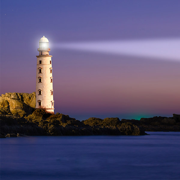 Lighthouse casting a beam of light across a rocky coastline at dusk