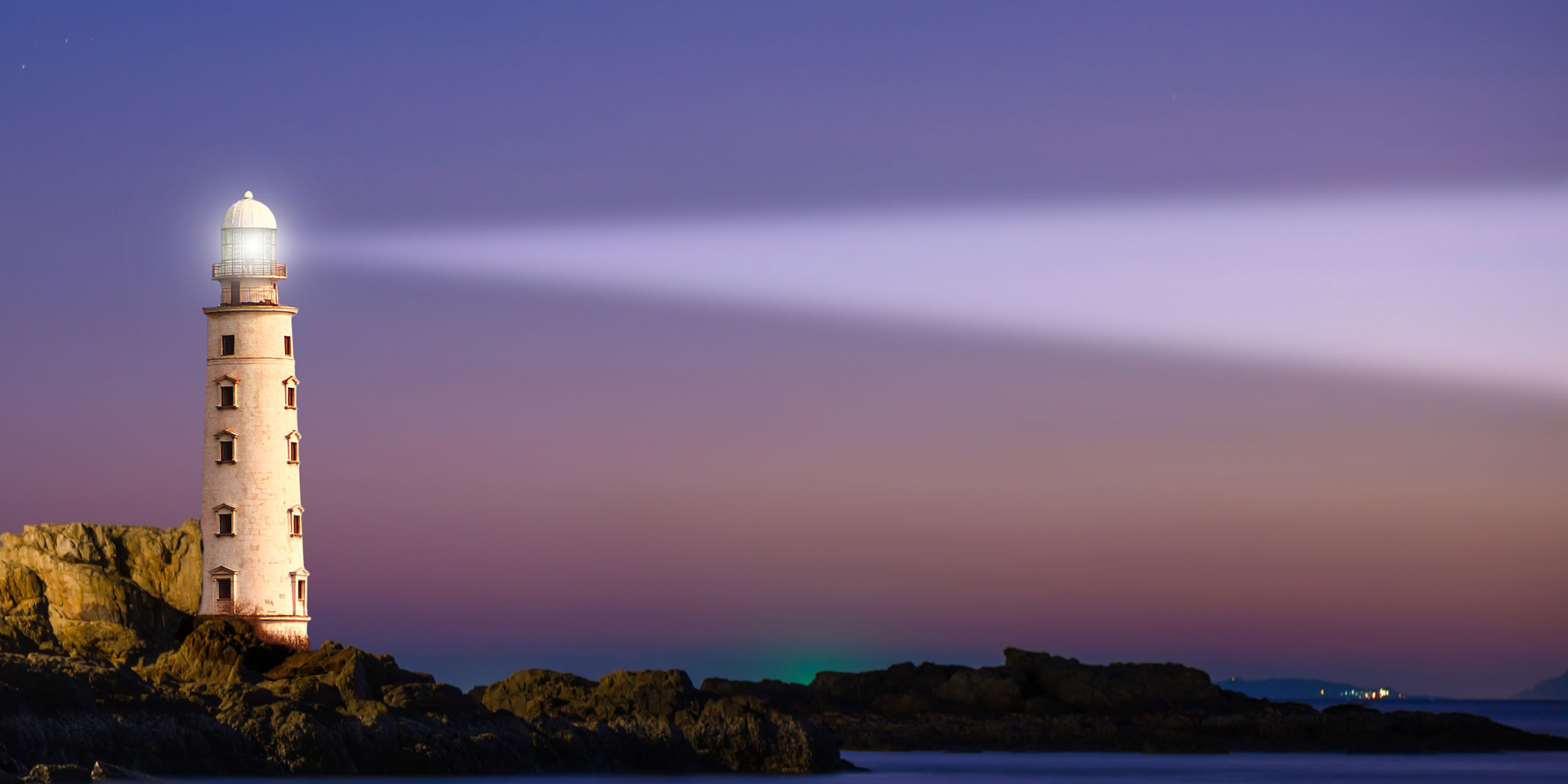 Lighthouse casting a beam of light across a rocky coastline at dusk